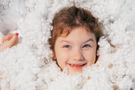 Close up smiling playful little girl with blue eyes in white soft feather bed, look at camera. Sweet dreams and cozy bedの写真素材