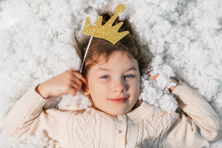 Close up calm, smiling little girl with blue eyes lying on soft cotton feather bed, hold toy crown and look at cameraの写真素材