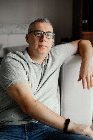 Portrait of peaceful middle-aged man sitting on sofa, leaning on hand at home, looking at camera, resting, relaxing.の写真素材