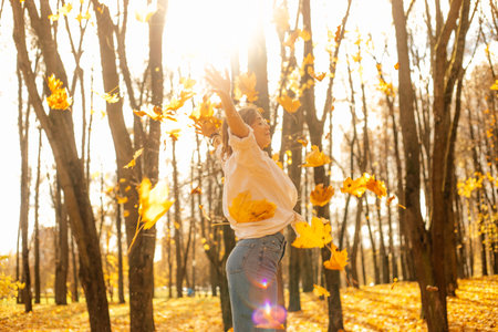 Side view of young cheerful teenage girl throwing yellow fallen maple leaves, stretching hands...
