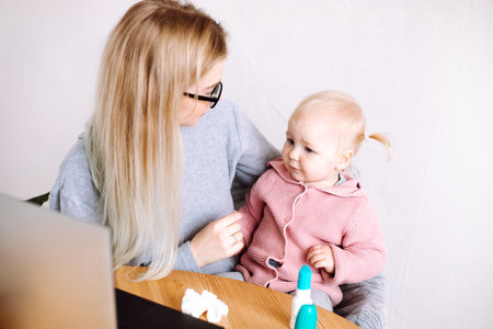 Portrait of young mother woman sitting at table near laptop, holding little worried baby. Working during sick time.の写真素材