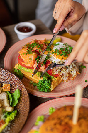 Vertical unrecognizable woman hands use knife, cut tasty, juicy sandwich pizza with omelette fried eggs on plate in cafeの写真素材
