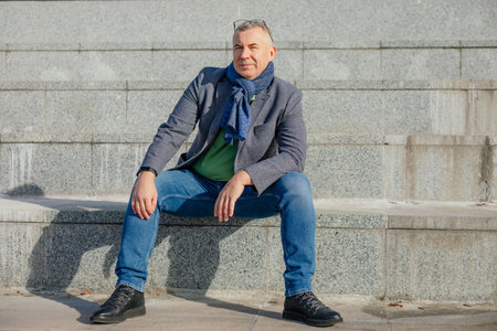Portrait of serious middle-aged man wearing grey jacket, warm blue scarf, glasses, jeans, sitting on...