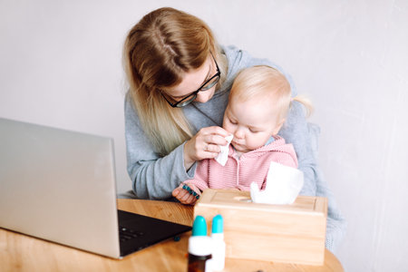 Portrait of young mother sitting at table near laptop, holding little baby, wiping running nose with napkin at home.の写真素材