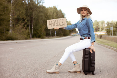 Nice woman try to stop passing car sitting on suitcase with outstretched cardboard poster on empty highway. Lady in hat escape from city to go anywhere. Travelling, hitchhiking, vacations, autumnの写真素材