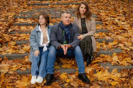 Happy family of three sit on stairs covered with fallen yellow leaves. Parents and teenage child...