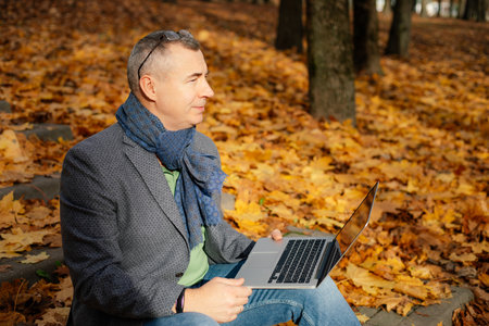 Aged handsome businessman sit on stairs covered with yellow fallen leaves in forest and work on laptop, side view.の写真素材