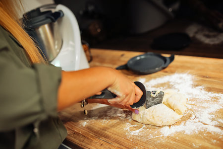 Hands of woman cutting kneaded dough into pieces by knife, selective focus. Cook on wooden countertop at home kitchen.の写真素材