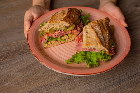 Hands of human hold plate of fresh appetizing sandwich on table background closeup, top view. Serving of sliced meat burger with greens, vegetables and crisp crust. Breakfast food, beautiful servingの写真素材