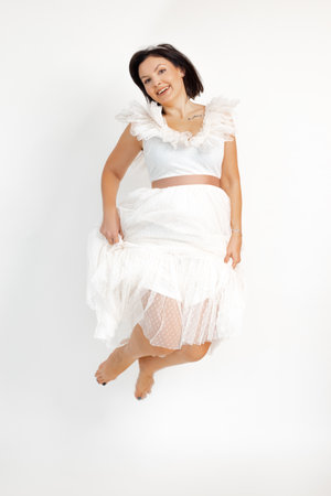 Bare footed happy adult brunette woman joyfully jump in airy romantic white dress, isolated on white background, copy space. Holiday look, event celebration, elegant female clothes, festive outfitの写真素材