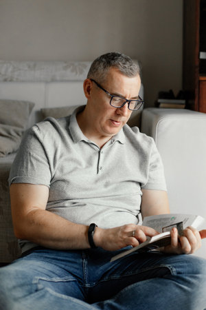 Vertical serious gray haired man in eyeglasses reading book, work in textbook, sitting on sofa...