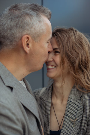 Vertical romantic couple of mature gray haired man kissing to nose fair haired laughing woman against gray wall. Familyの写真素材