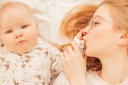Cheerful young mother kiss fingers on palm of little baby lying on bed closeup. Home family photo of...