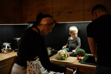 Parents with little child cook dinner in kitchen with dark light. Prepare fresh cauliflower head for cutting on board.の写真素材