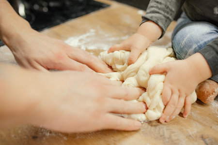 Parent with baby together knead raw elastic dough on kitchen tabletop closeup. Little hands help mom cooking dinner.の写真素材
