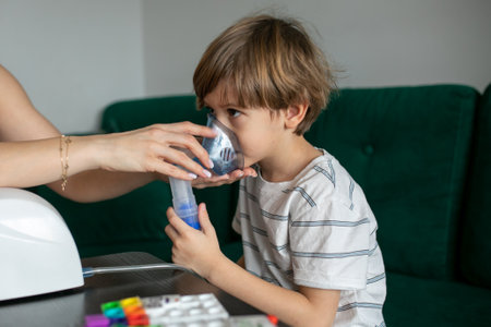 Woman inhaling child at home with inhaler. Sick little boy sit on sofa. Breathing vapor, respiratory disease, bronchitisの写真素材