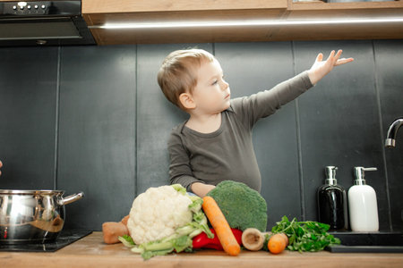 Little child helping cook dinner. Baby son sitting with raised arm on tabletop among pile of fresh ripe vegetables.の写真素材
