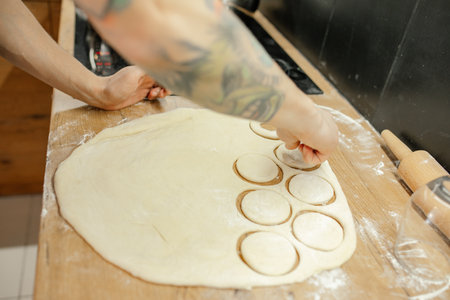 Male hands cut out round pieces with glass from raw thin dough. Roll out dough with rolling pin on...