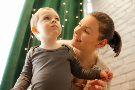 Happy mother and little boy embracing, from below view. Family home portrait. Garland on curtains decorate interior.の写真素材