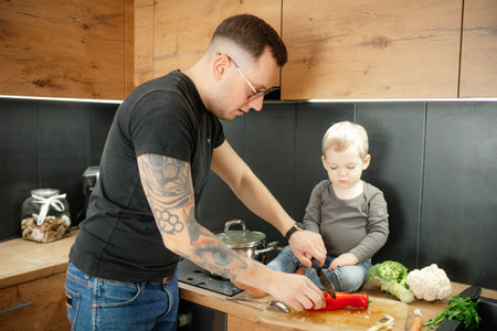 Father with little child cook dinner side view. Baby son sitting on tabletop help dad cut red bell pepper on board.の写真素材