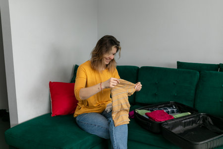 Portrait of smiling exciting young woman sitting on green sofa near suitcase at home, packing...