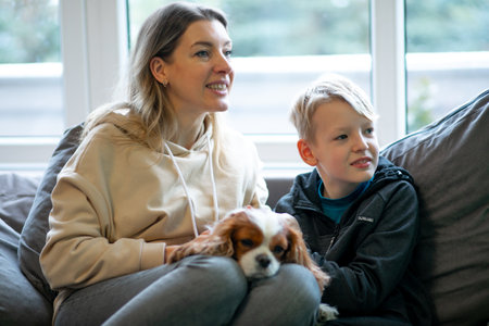Mother and son sitting on sofa near window with spaniel. Family portrait in cozy home. Sad puppy lying on female legs.の写真素材
