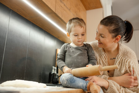Tender laughing positive brunette woman carrying little blond boy with rolling pin in hands, cooking...