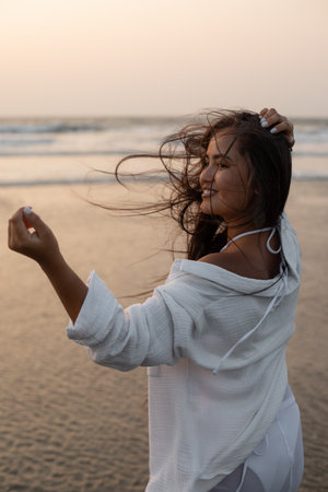 Young beautiful smiling brunette with loose long hair is standing turned around on seashore against...