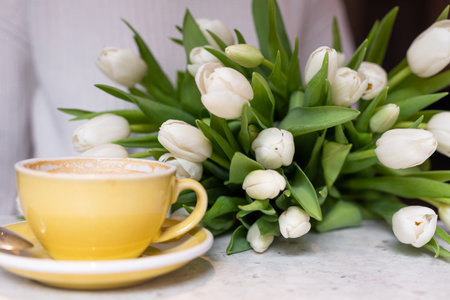 Yellow ceramic mug with cappuccino is on table in cafe, next to it lies luxurious bouquet of tulips. Floral surprises.の写真素材