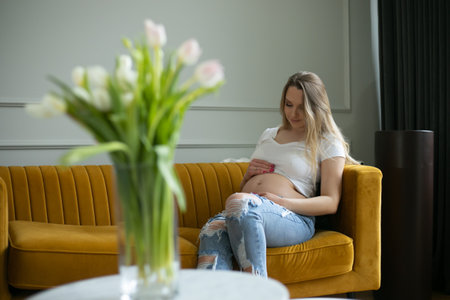 Happy pregnant woman touch with hands bare belly sitting on sofa in living room. Blurry bouquet of white tulips on tableの写真素材