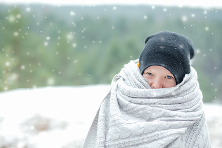 Funny frozen little boy in warm hat, wrapped in wool blanket and look at camera against snowy winter forest. Copy spaceの写真素材