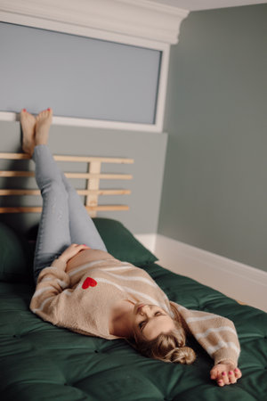 Happy pregnant woman in home clothes lie on bed with raised legs. Modern bedroom interior design, resting place.の写真素材
