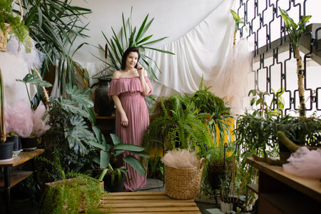 Organization of home greenhouse. Charming female florist in pink off-the-shoulder dress stands in her greenhouse.の写真素材