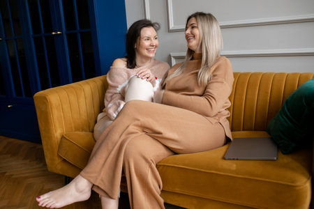 Beautiful women in textile suits relax on stylish mustard color sofa and joyfully talk in living room. Friends meeting.の写真素材
