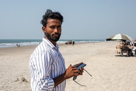 Charming Indian man is standing on beach against background of sea, holding phone in his hands and looking at camera.の写真素材