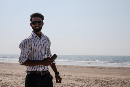 Attractive Indian man stands on beach , holding phone in his hands, smiling and looking at camera. Communications.の写真素材