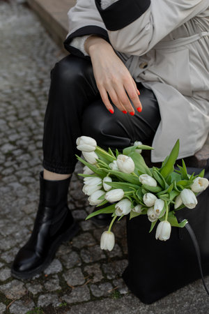 Young woman in light coat, sat down to rest on city street, next to black bag with bouquet of tulips. Stone pavement.の写真素材