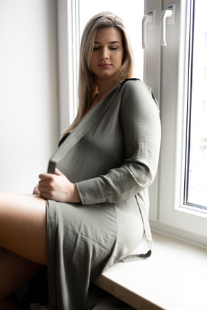 Portrait of attractive long-haired pregnant woman in gray long shirt, who is sitting on windowsill opposite window.の写真素材