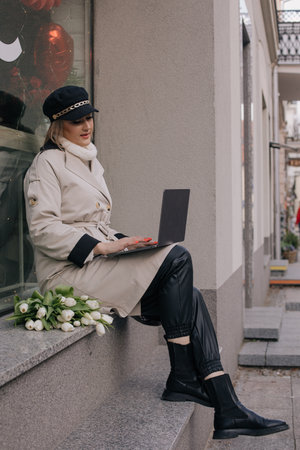 Modern street fashion. Stylishly dressed woman with laptop on her lap is sitting on city street , next to bouquet.の写真素材