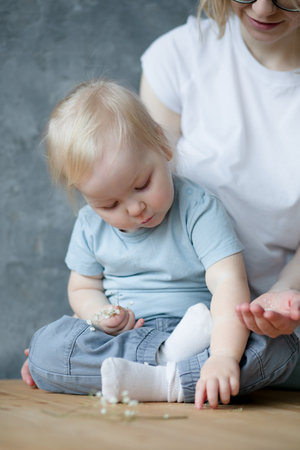 Young woman mother holding tiny white flowers for little blond girl baby. Daughter sitting gathering flowers from table.の写真素材