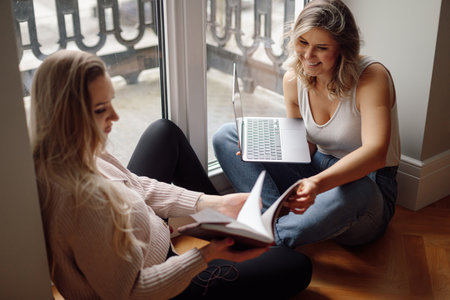 Portrait of cheerful young women holding grey laptop, looking at open book, debating, talking, coworking. Technology.の写真素材