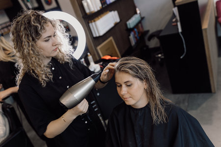 Young woman with long hair sit in beauty salon. Hairdresser making hairstyle using dryer near ring lamp. Beauty blogger.の写真素材