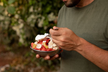 Cropped photo of young man holding tropical fruit salad with cut various exotic fruits metal bowl, fork, eating outside.の写真素材