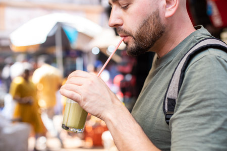 Side view of young bearded man wearing khaki T-shirt, standing on local market, holding drinking glass with cocktail.の写真素材
