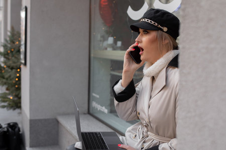 Portrait of woman in black cap with laptop in hands talking on phone from window of house looking urban viewの写真素材