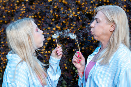Mother and daughter close eyes and blowing on dandelions. Relatives make wish, have fun and enjoying...