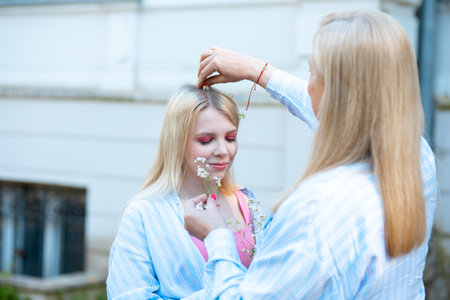 Mother help daughter decorate hair with little white flowers. Girl close eyes and trust mom. Plants in blondie hair.の写真素材