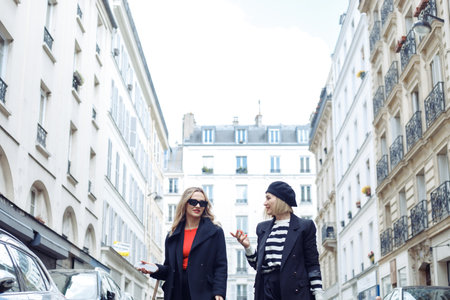 Two women walk together on street in romantic Paris. Adult blondes in black outfits, with red lips...