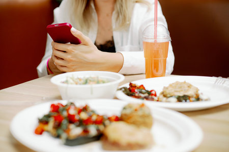 Young woman freelancer sitting at table with dishes in cafe and holding phone. Concentrated lady working at lunch time.の写真素材