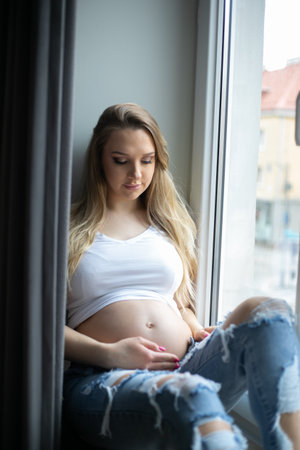 Glorious gravid woman comfortable resting on windowsill behind black curtains at home and gently touching bare belly.の写真素材